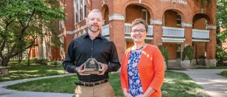 Two people posing for a picture outside of a brick building holding an award.